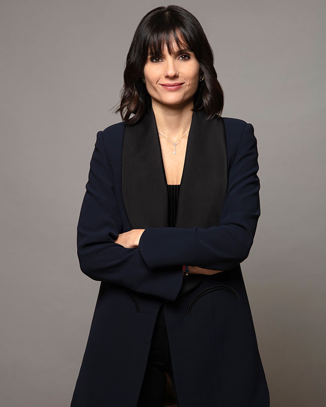 Portrait de la journaliste libanaise Dalal Mawad, en manteau bleu nuit, les bras croisés, sur fond neutre